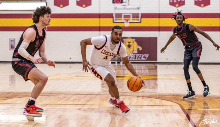 A basketball player in a white 'Cougars' jersey dribbles past two defenders in black on a gym court.